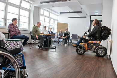 In einem Raum sitzen einige Menschen unter anderem in Rollstühlen in verschiedenen Altersgruppen