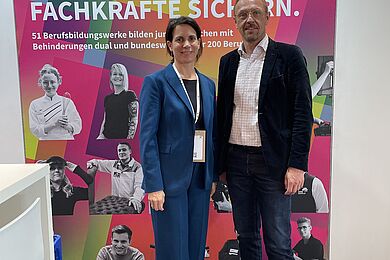 Fotowand der BAG BBW, davor Tanja Ergin, Geschäftsführerin BAG BBW (links) in einem blauen Hosenanzug, rechts Tobias Schmidt, Vorstandsvorsitzende*r BAG BBW mit weißem Hemd, dunklem Jackett und Jeans.