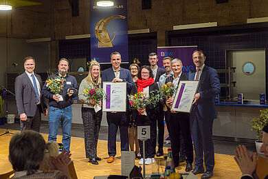 Gruppenfoto nach der Preisvergabe der Brückenschlag-Preise. Alle Preisträger*innen und die Geschäftsführung der BBW-Leipzig-Gruppe mit Blumensträußen, den Preisen und den Urkunden auf der Bühne.