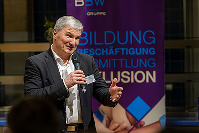 Steffen Leonhardi, Vorsitzender der Geschäftsführung der Agentur für Arbeit Leipzig, hält in der rechten Hand ein Mikrofon. Mit der linken Hand gestikuliert er. er scheint jemanden im Publikum anzusprechen. 